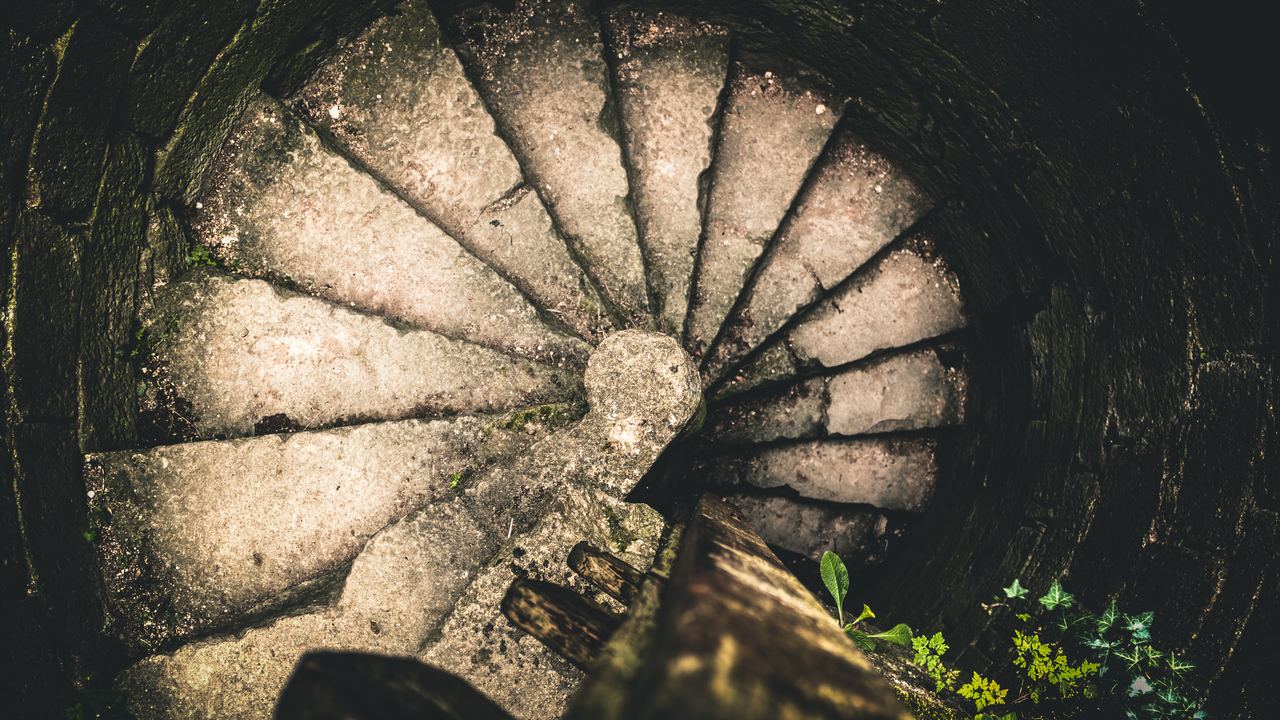 A worn stone spiral staircase in a castle seen from directly above, its wedge-shaped treads turning around the central newel post.
