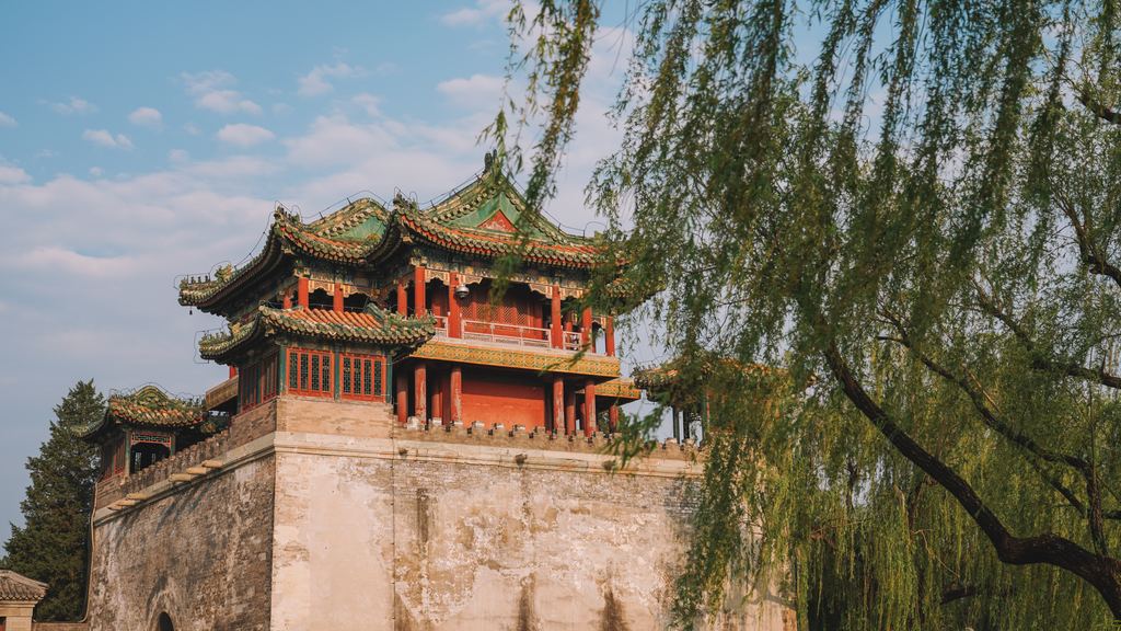 A painted timber pavilion on a stone terrace in the Summer Palace, framed by the hanging branches of a willow.