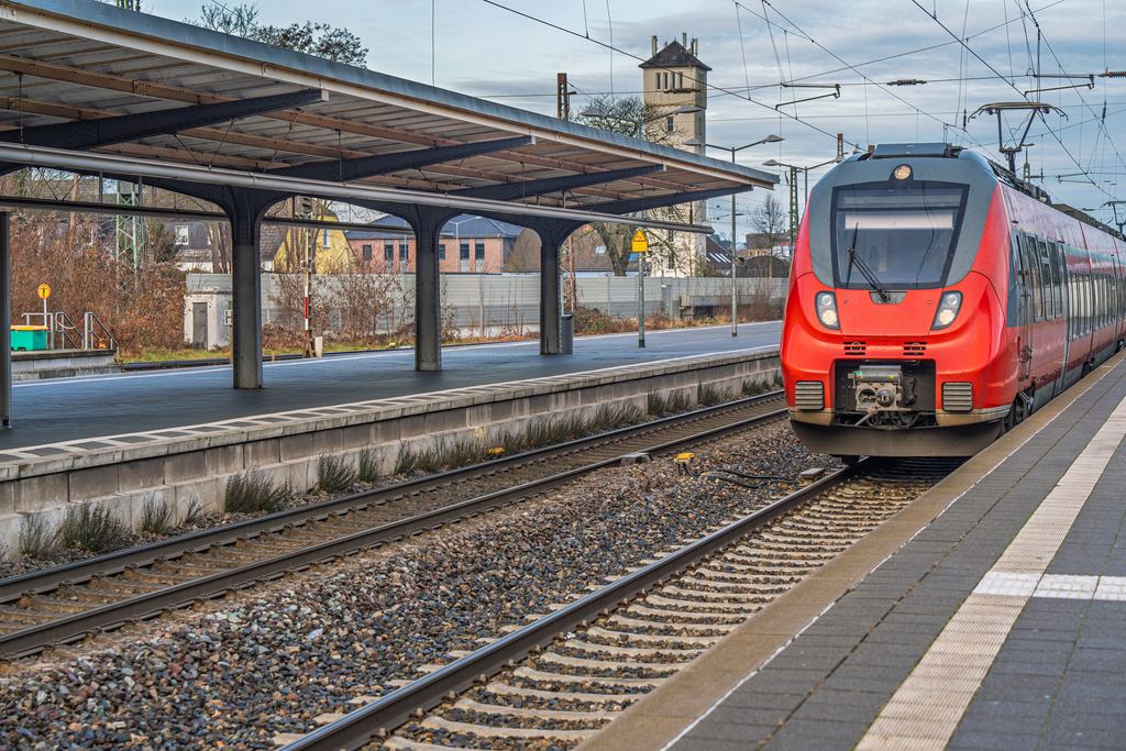 A red regional train drawing into a small German station platform, with an old tower visible above the canopy roof.