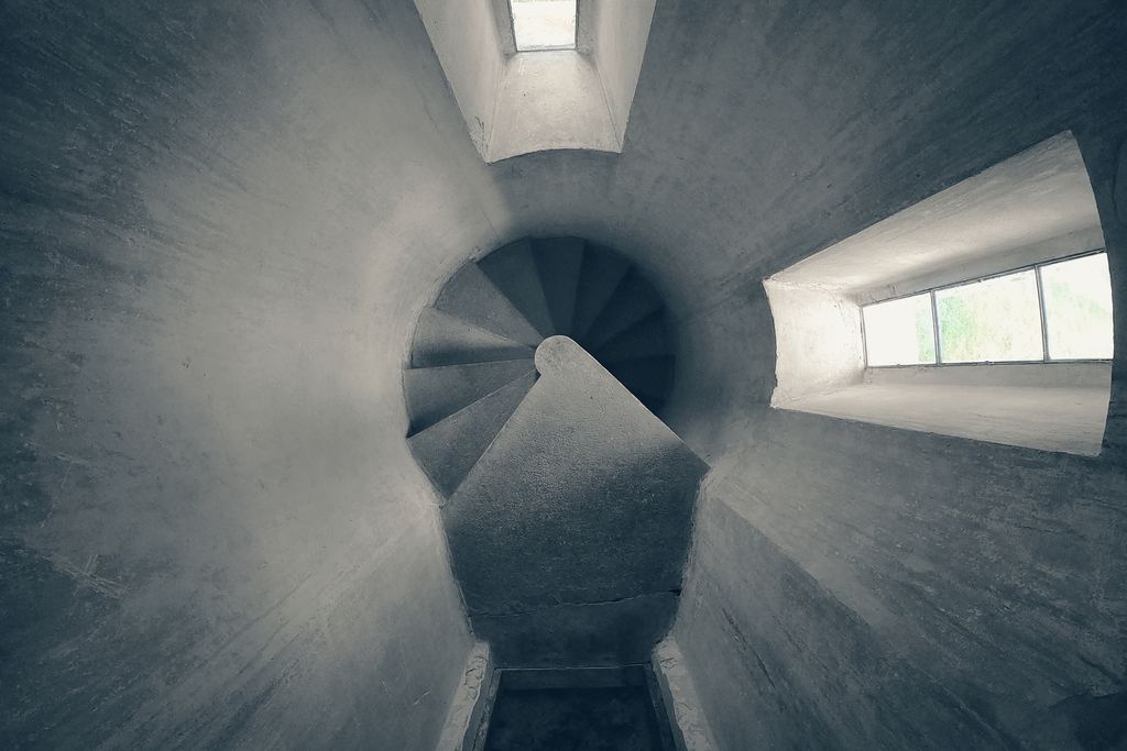 Looking down a grey stone stair tower in a fortress, with narrow slit windows cut through the wall at each turn.