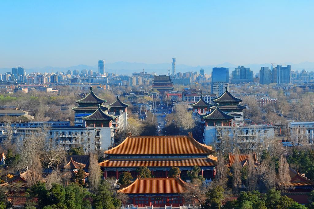 The tiled roofs and painted eaves of old Beijing in the foreground, with the towers of the modern city rising behind them.