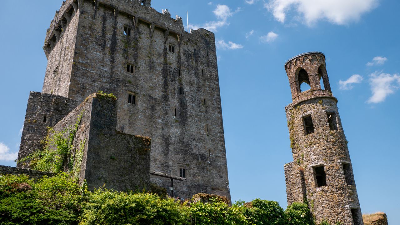 The battlemented keep of Blarney Castle and the ruined round tower beside it, seen from below against a blue sky.