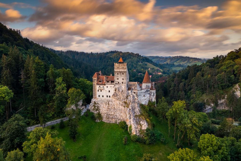 Bran Castle standing on its rock above the forested Carpathian foothills, its walls lit gold under a sky of broken cloud.