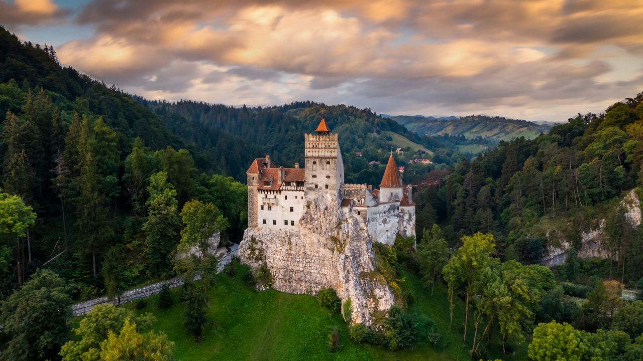 Bran Castle standing on its rock above the forested Carpathian foothills, its walls lit gold under a sky of broken cloud.