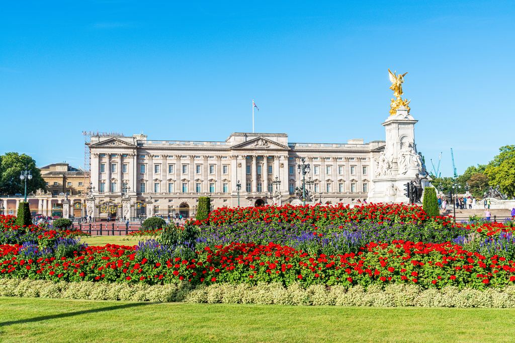 The east front of Buckingham Palace behind beds of red flowers, with the gilded Victoria Memorial on the right.