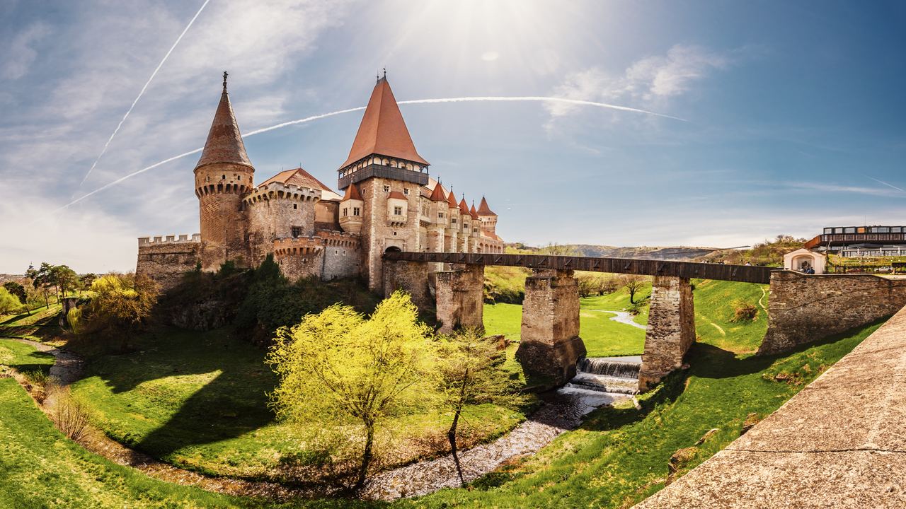 The red-roofed towers of Corvin Castle in Hunedoara with the long wooden bridge running to its gate over the green ravine.