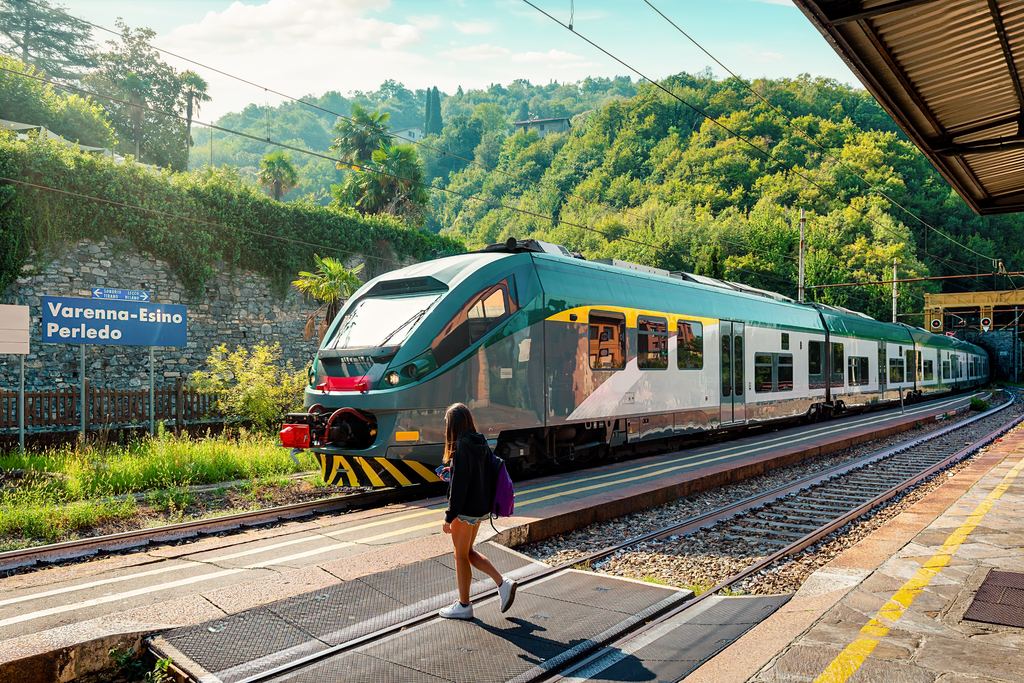 A traveller stepping off the platform of a small Italian country railway station, with a regional train alongside and wooded hills behind.