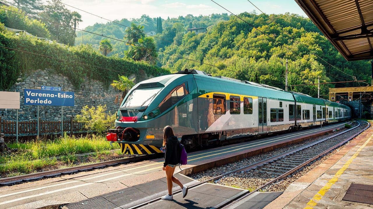 A traveller stepping off the platform of a small Italian country railway station, with a regional train alongside and wooded hills behind.