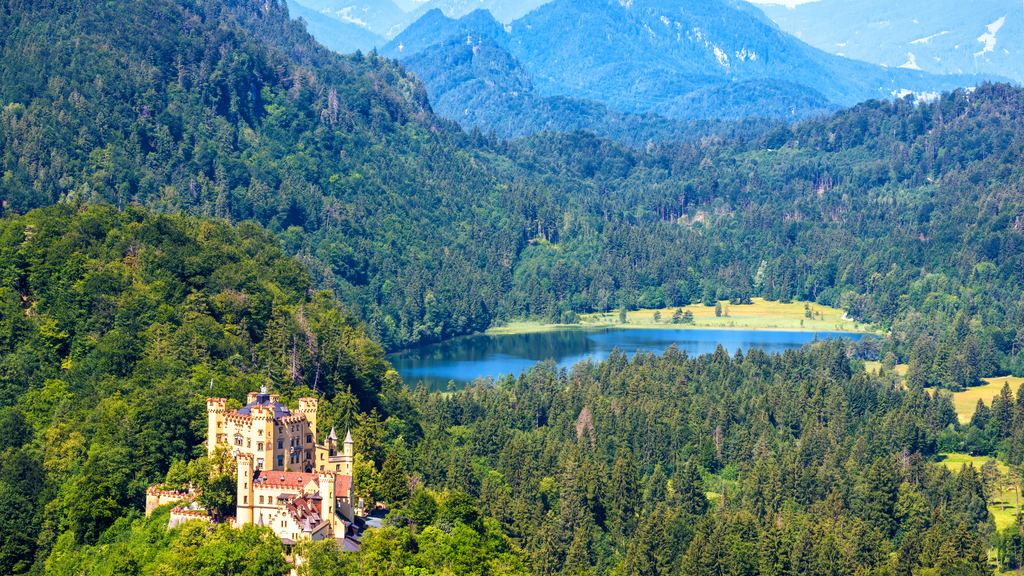 The yellow walls of Hohenschwangau Castle under the wooded slopes of the Bavarian Alps, with a lake in the valley beyond.