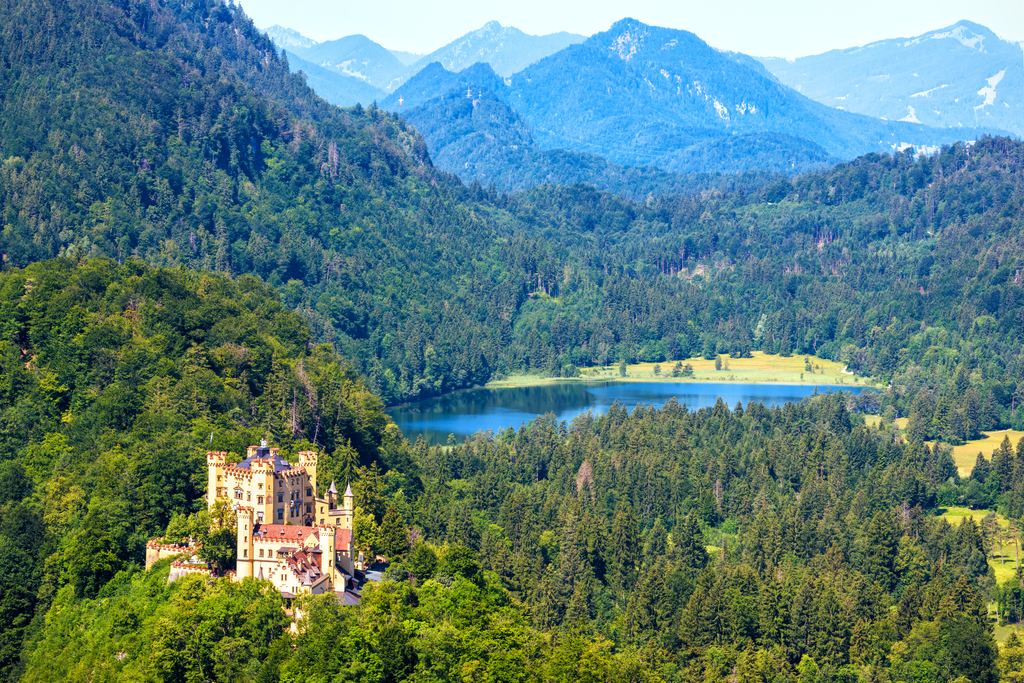 The yellow walls of Hohenschwangau Castle below the wooded slopes of the Bavarian Alps, with a lake in the valley beyond.