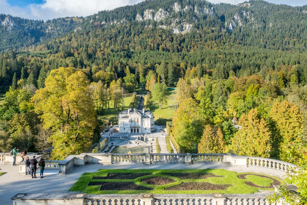 The white Linderhof Palace and its formal parterre set in a forested Bavarian valley, with wooded ridges rising behind.