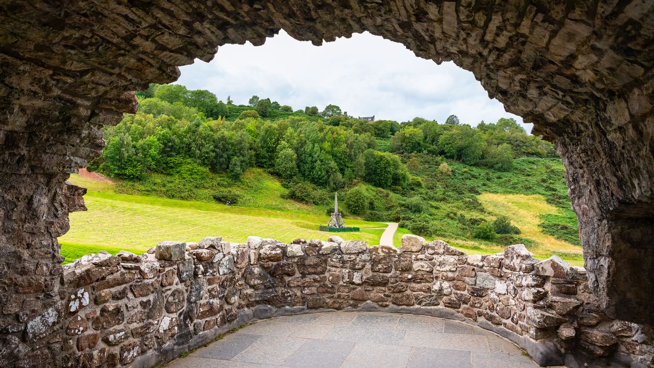 A broken stone arch at Urquhart Castle framing the green banks of Loch Ness, with nothing but grass and sky where rooms once stood.