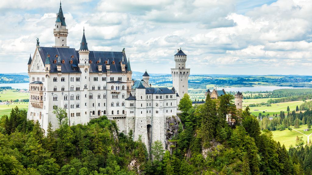 Neuschwanstein Castle standing on its wooded spur with its white towers and blue turrets, the Bavarian plain stretching behind.