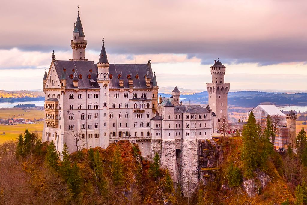 Neuschwanstein Castle at sunset, its towers and gatehouse warm against a pink sky above autumn-coloured forest.