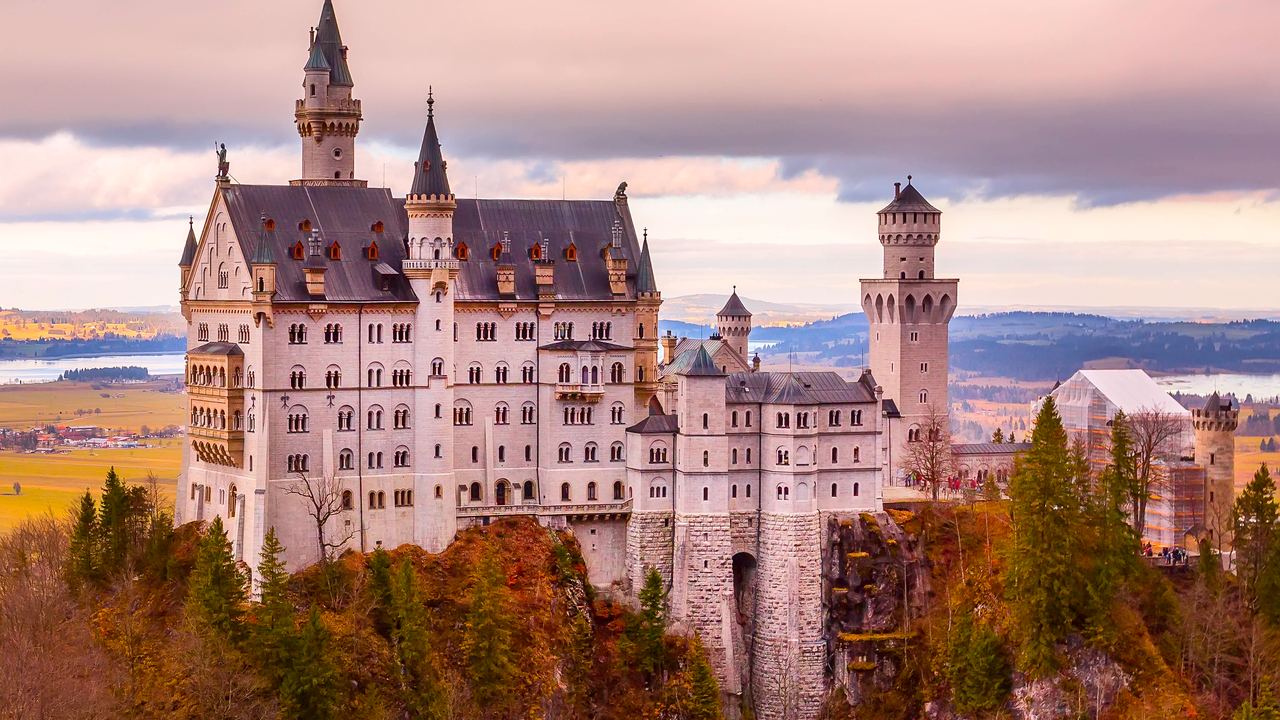 Neuschwanstein Castle at sunset, its towers and gatehouse warm against a pink sky above autumn-coloured forest.
