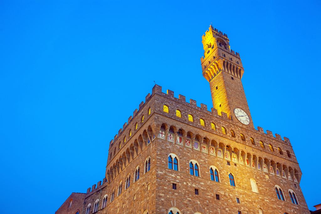 The rusticated front and lit bell tower of Palazzo Vecchio in Florence standing against a deep blue twilight sky.