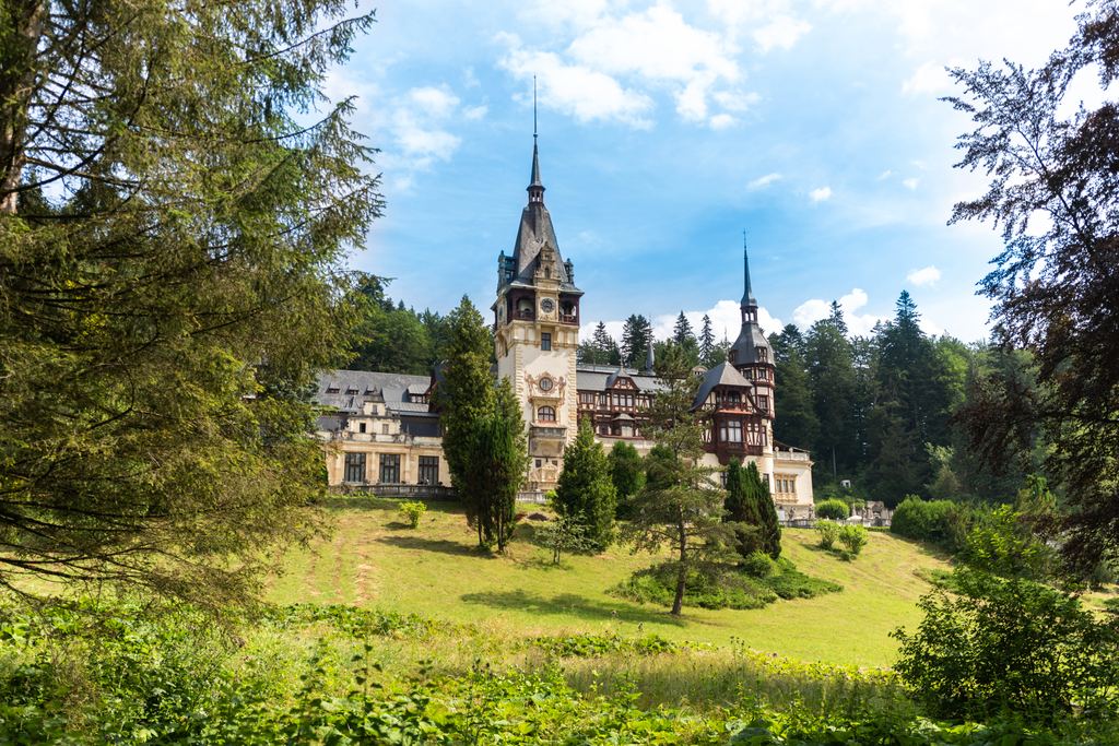 Peles Castle at Sinaia seen across a green lawn and framed by tall conifers, its spires and timbered gables against the sky.