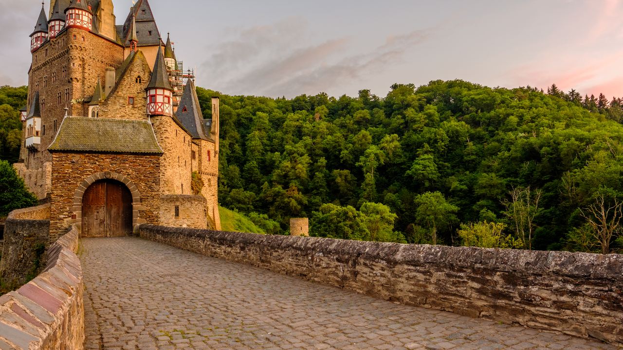 The cobbled approach and gate tower of Burg Eltz, its walls warm in evening light above the wooded slope.