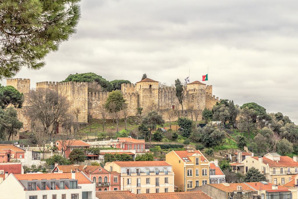 The Castelo de Sao Jorge on its hill above the roofs of Lisbon, its curtain wall and towers under a broken sky.