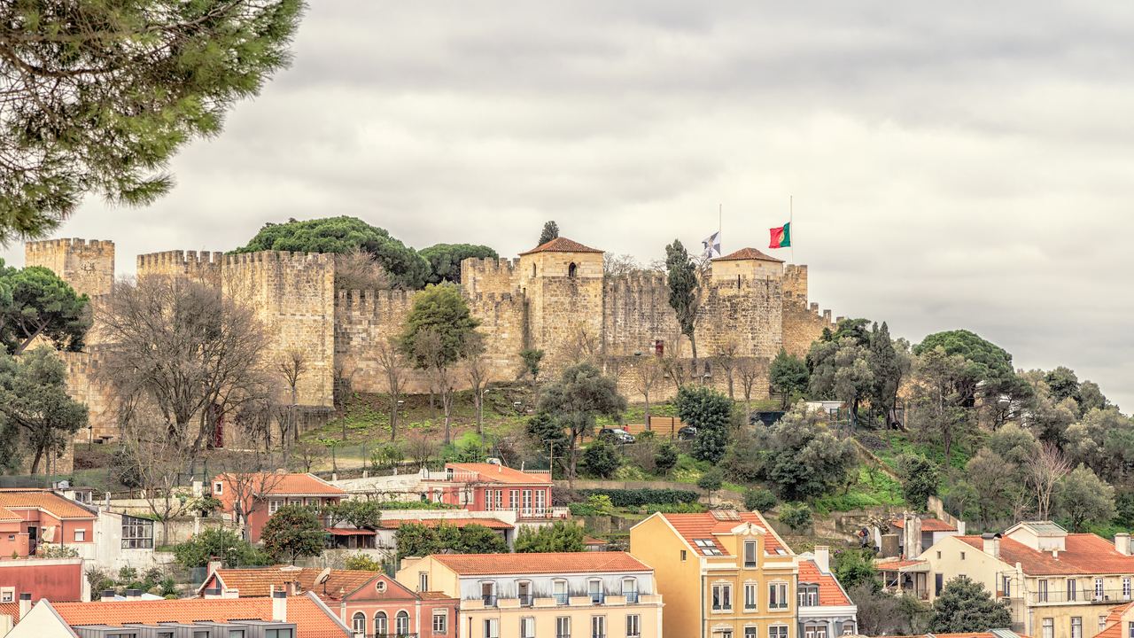 The Castelo de Sao Jorge on its hill above the roofs of Lisbon, its curtain wall and towers under a broken sky.