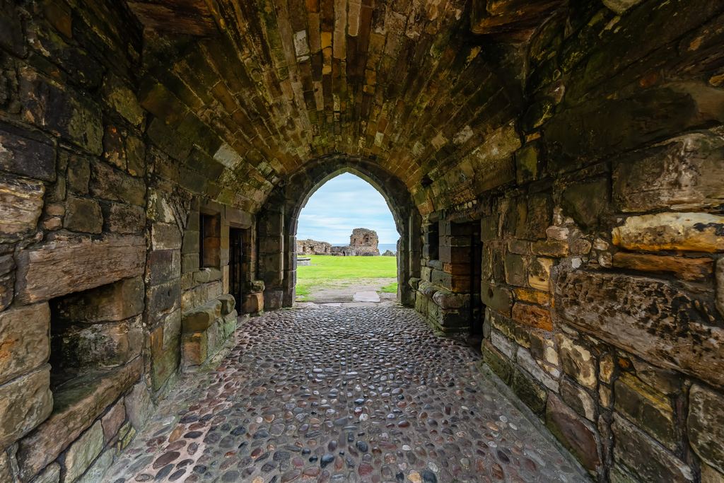 A vaulted stone entrance passage at St Andrews Castle paved with rounded cobbles, opening onto the grass and ruins beyond.