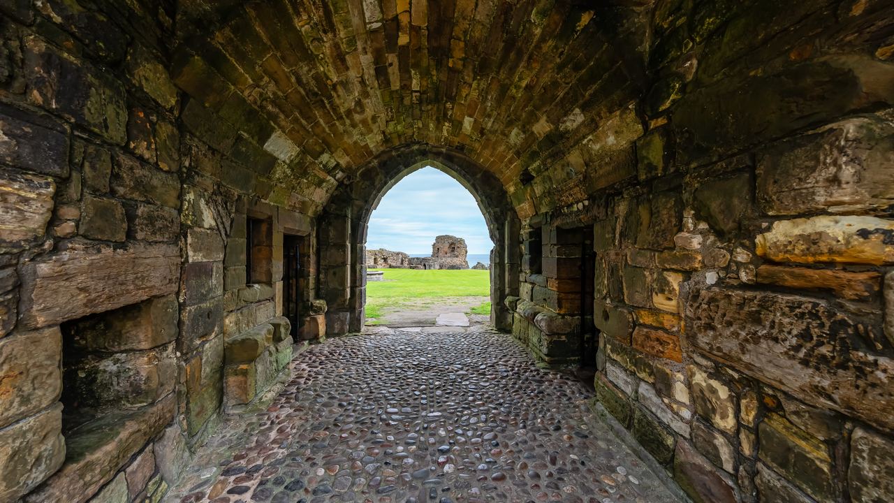 A vaulted stone entrance passage at St Andrews Castle paved with rounded cobbles, opening onto the grass and ruins beyond.