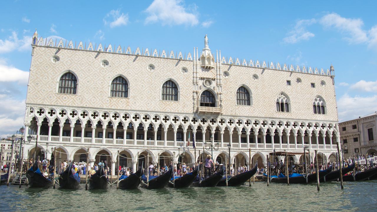 The full pink and white front of the Doge's Palace along the Venetian waterfront, with gondolas moored in front of it.