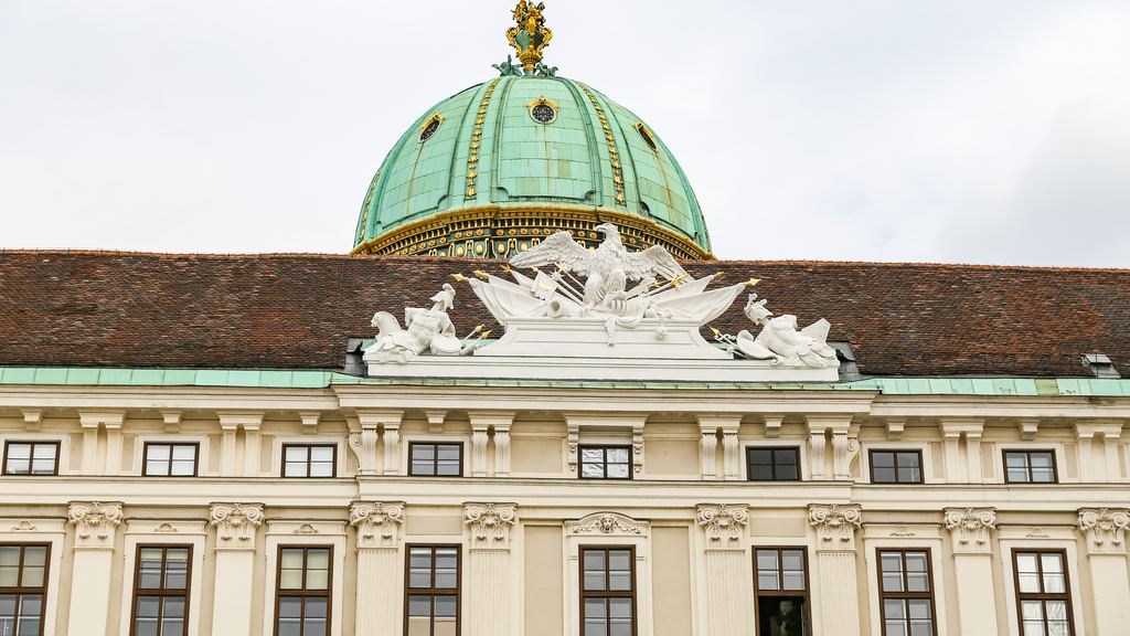 The green copper dome and carved stone figures of the Hofburg palace in Vienna standing against a pale sky.
