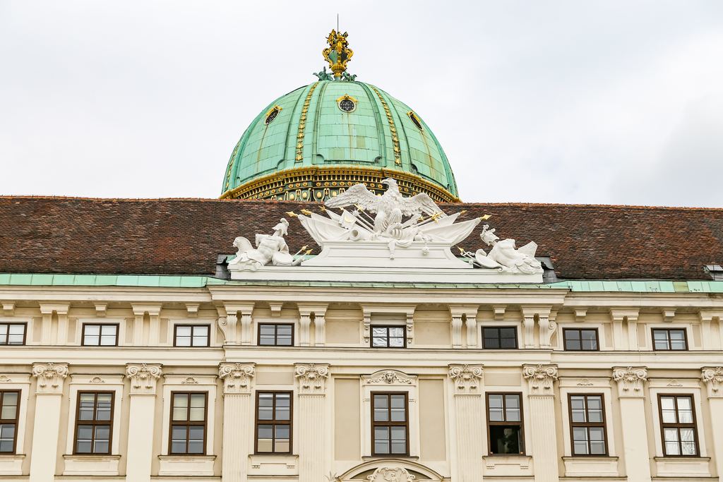 The green copper dome and carved stone figures of the Hofburg palace in Vienna standing against a pale sky.