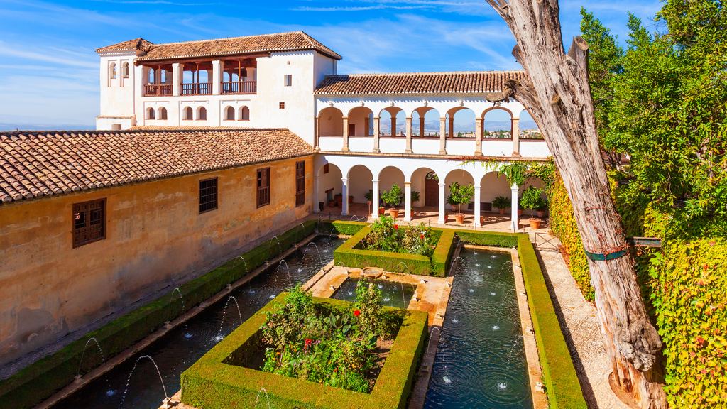 The whitewashed pavilion of the Generalife above the long water channel and clipped hedges of its garden, under a bright sky.