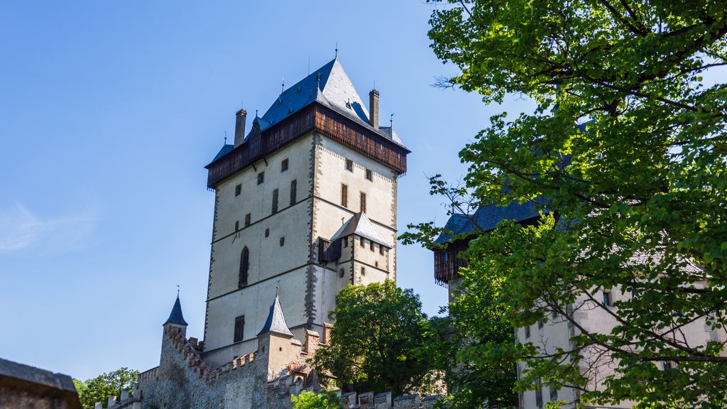 The white Great Tower of Karlstejn Castle with its dark roof rising above the trees against a clear blue sky.