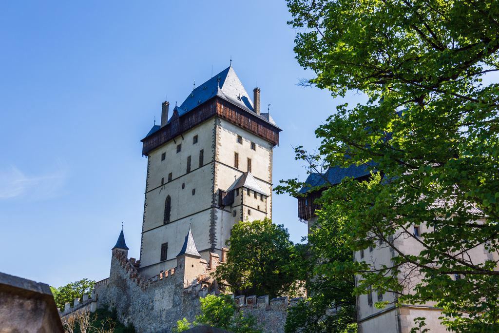 The white Great Tower of Karlstejn Castle with its dark roof rising above the trees against a clear blue sky.