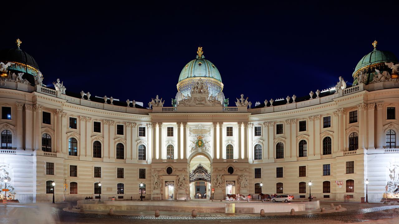 The curved wing and green dome of the Hofburg palace lit at night above Michaelerplatz in the centre of Vienna.