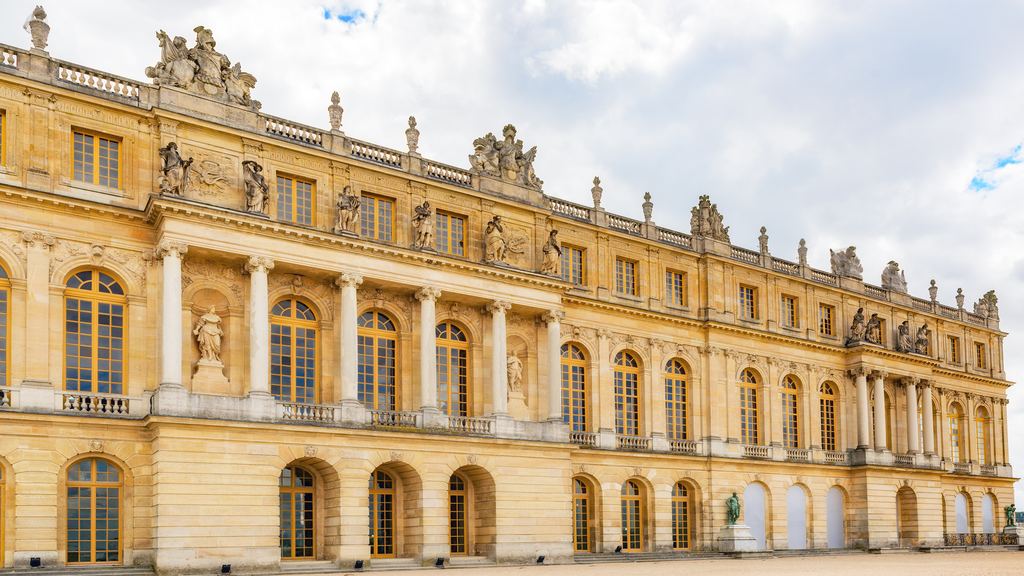 The gilded stone facade around the Marble Court at the Palace of Versailles under a cloudy sky.