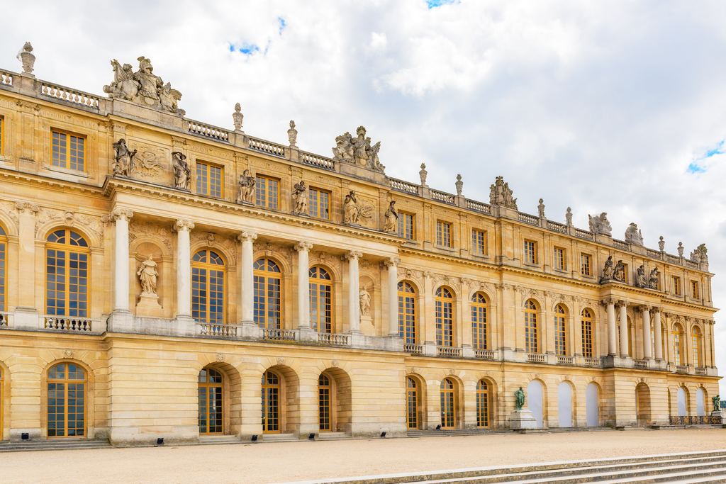 The gilded stone facade around the Marble Court at the Palace of Versailles under a cloudy sky.