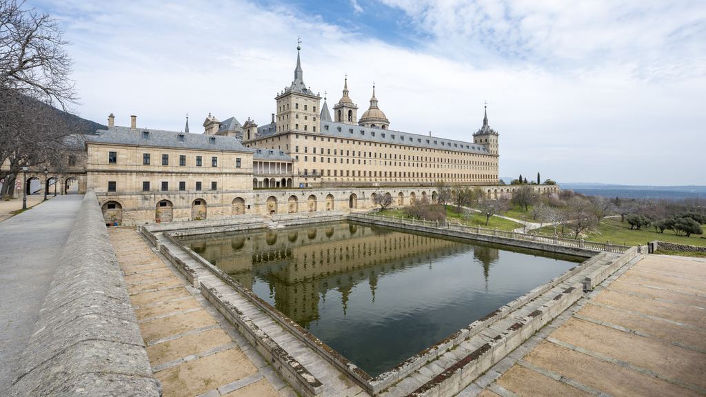 The granite front of the monastery of El Escorial reflected in the rectangular pond below it.