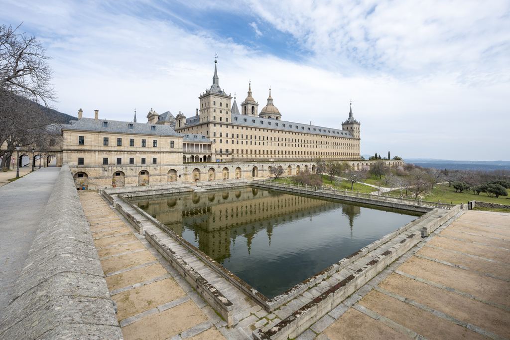 The granite front of the monastery of El Escorial reflected in the rectangular pond below it.