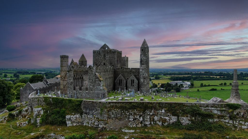 The cathedral and round tower of the Rock of Cashel standing in silhouette on their limestone outcrop under a pink evening sky.