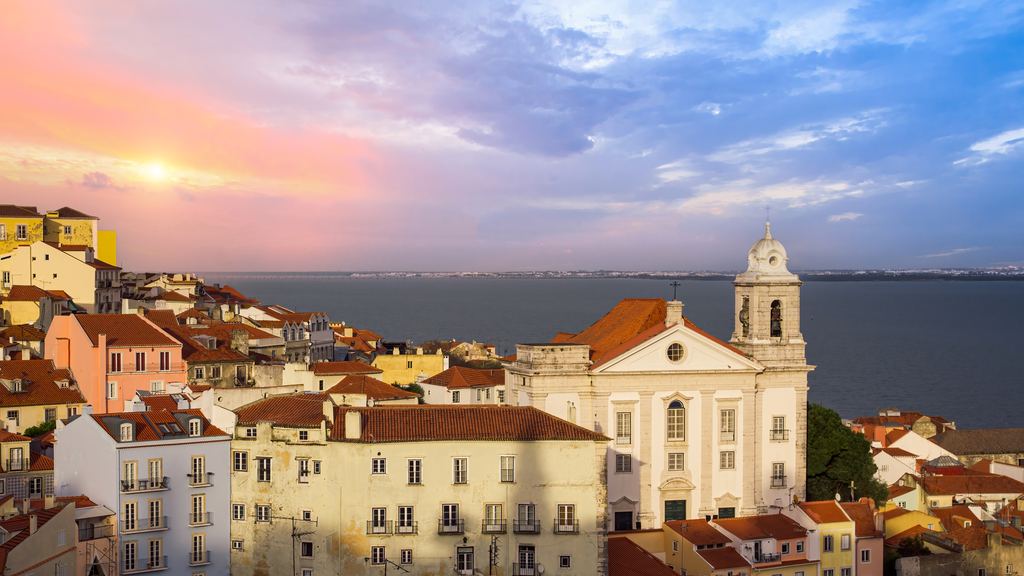 The roofs and church domes of the Alfama quarter of Lisbon under a sunset sky, with the Tagus estuary beyond.