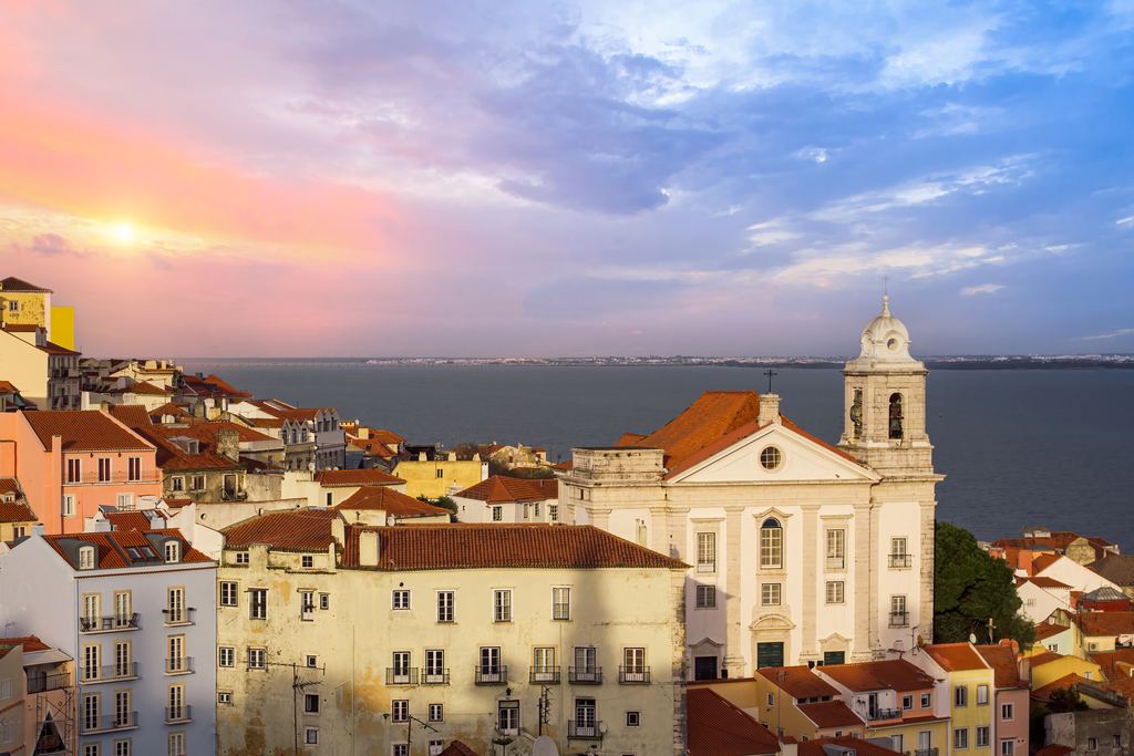 The roofs and church domes of the Alfama quarter of Lisbon under a sunset sky, with the Tagus estuary beyond.