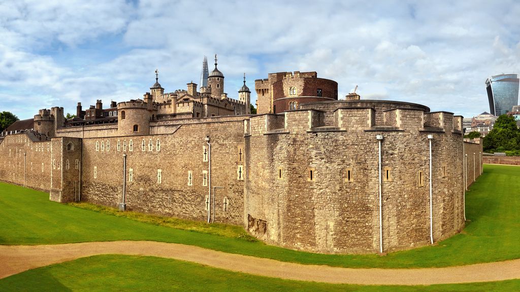 The outer curtain wall of the Tower of London seen across the grassed dry moat on a clear day.