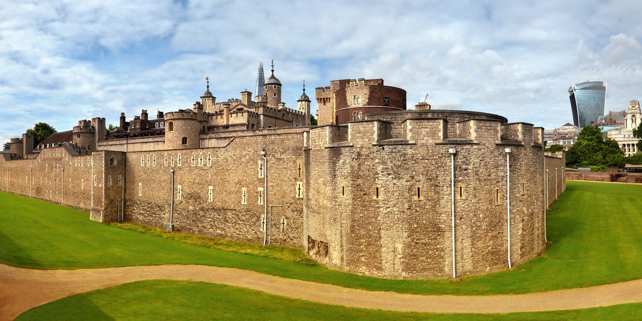 The outer curtain wall of the Tower of London seen across the grassed dry moat on a clear day.