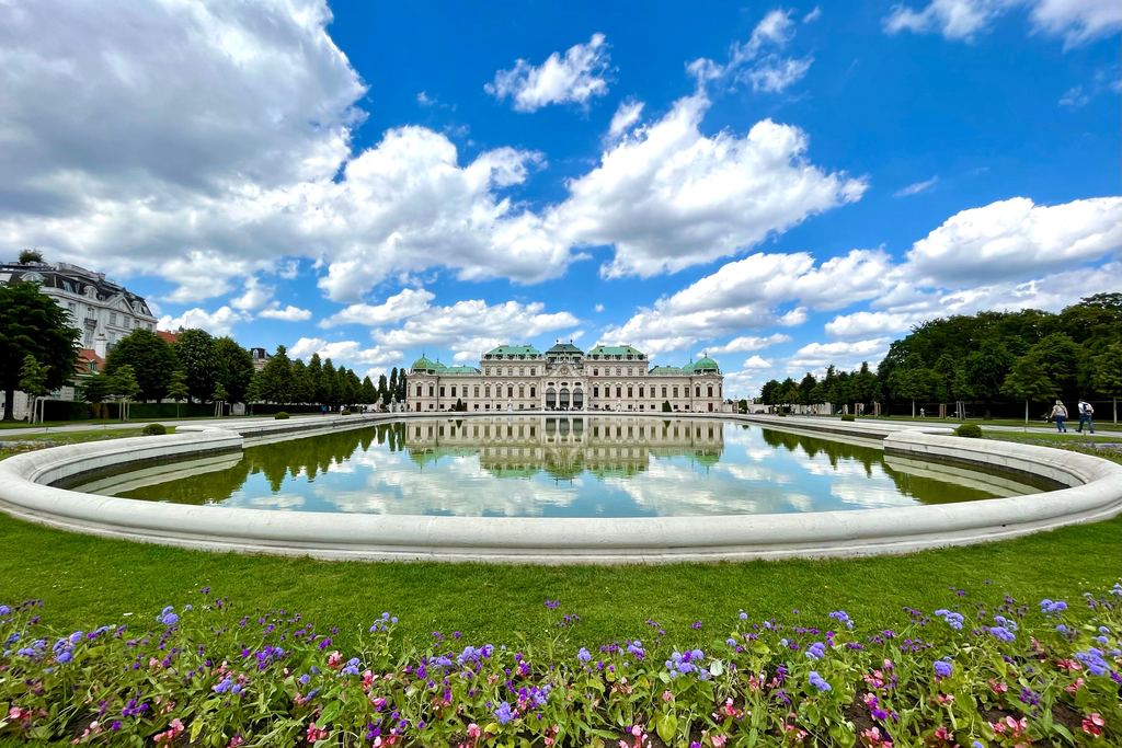 The Upper Belvedere palace in Vienna mirrored in its long reflecting pool, with beds of flowers under a sky of white cloud.