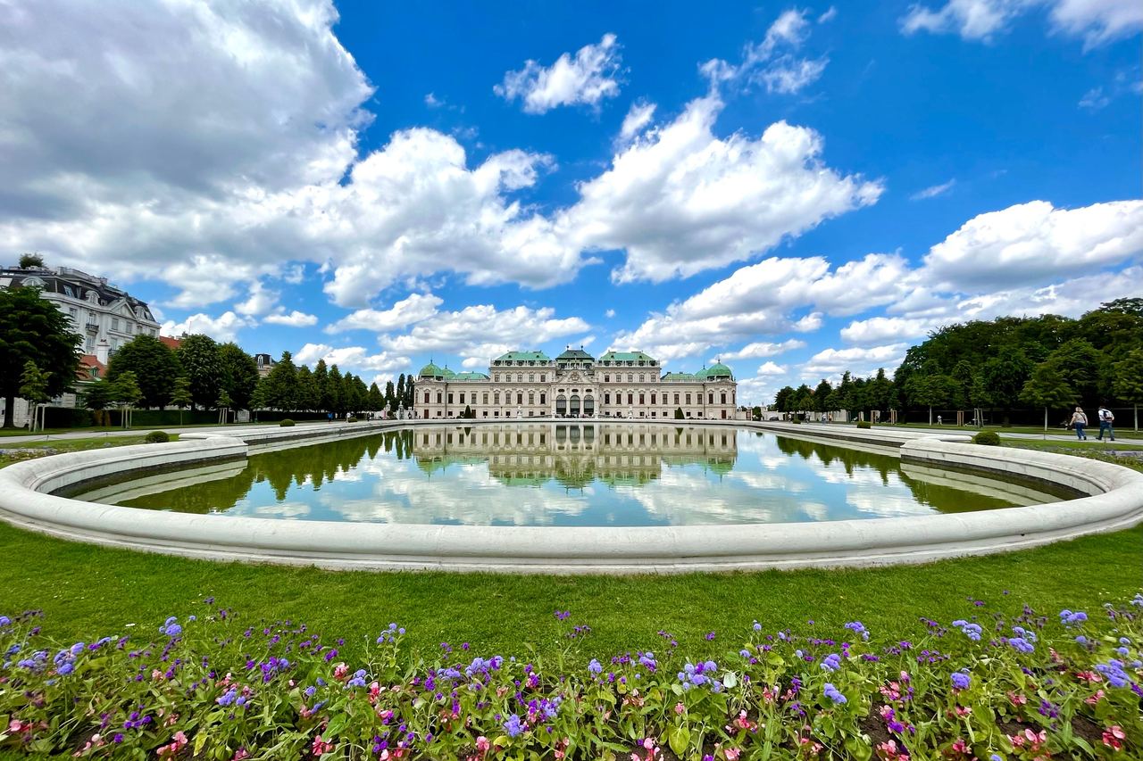 The Upper Belvedere palace in Vienna mirrored in its long reflecting pool, with beds of flowers under a sky of white cloud.