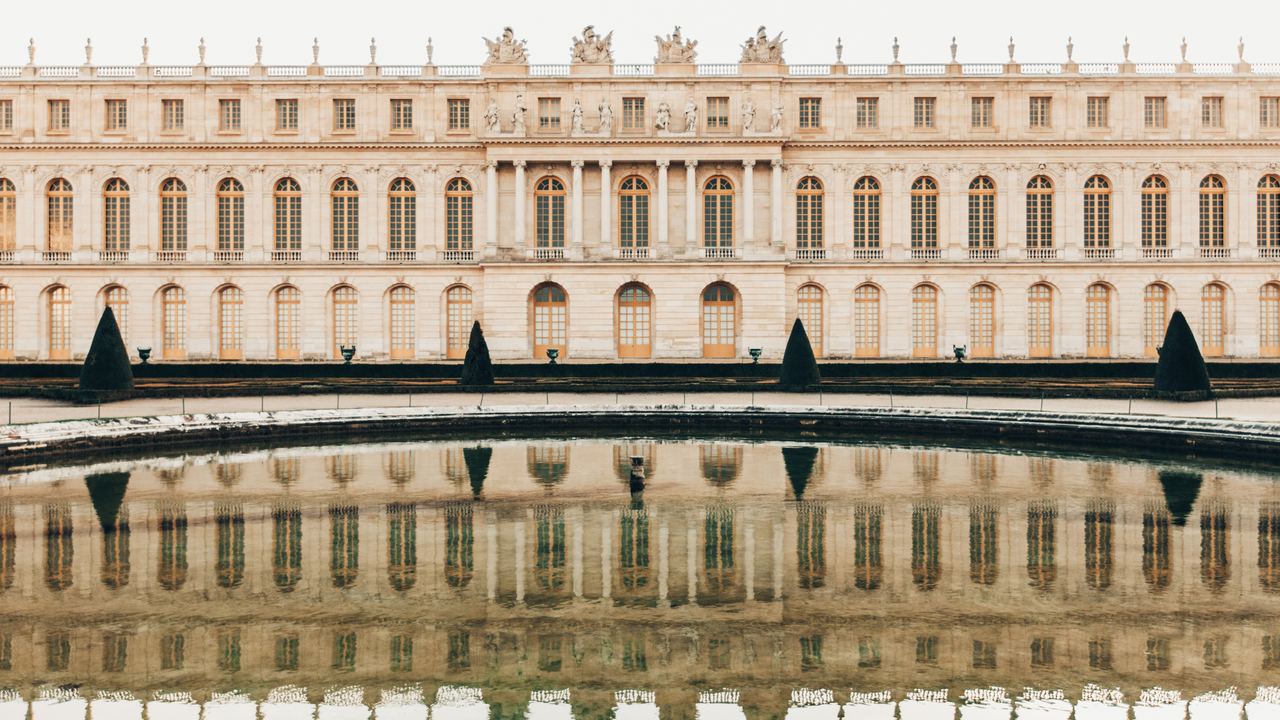 The long garden facade of the Palace of Versailles reflected in the still water in front of it.