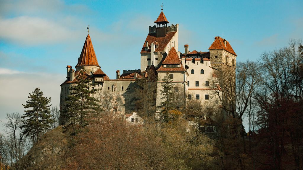 The white walls, red roofs and pointed tower of Bran Castle rising above the conifers of its Transylvanian hillside.