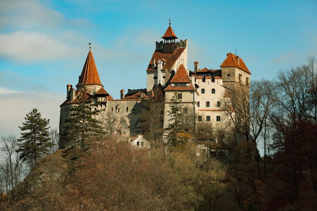 The white walls, red roofs and pointed tower of Bran Castle rising above the conifers of its Transylvanian hillside.
